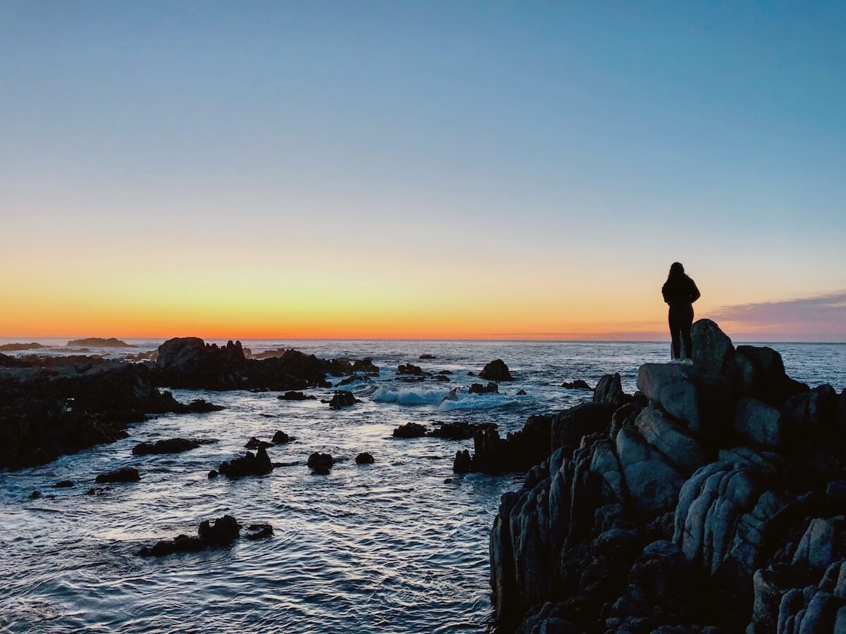 Elaina standing on the rocks by the ocean in Monterey focusing on the mental health benefits of the ocean.