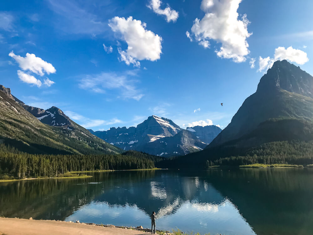 Mountains reflected in a lake in Glacier National Park in Montana with a man standing next to the lake