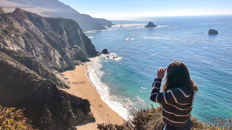 Elaina standing near the edge of a cliff at the outlook for the Bixby Creek Bridge in Big Sur. Elaina is looking out over the beach with the ocean in the background