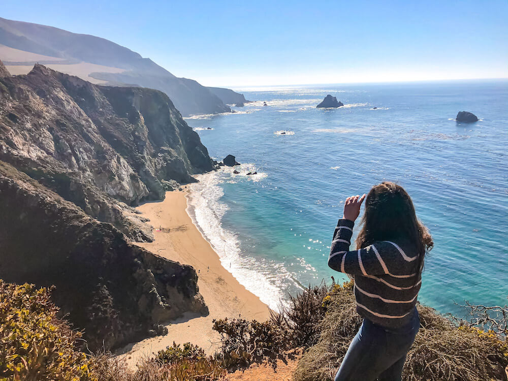 Elaina standing near the edge of a cliff at the outlook for the Bixby Creek Bridge in Big Sur. Elaina is looking out over the beach with the ocean in the background