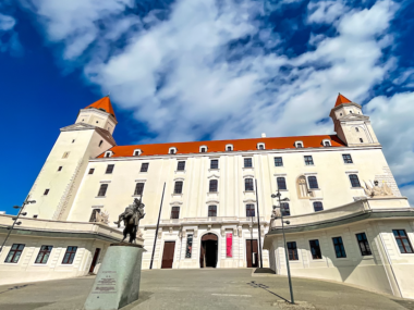 The front of Bratislava castle on a stop during one day in Bratislava. The building is white with a red roof and tower on either side. There is a statue in front of a man on a horse.