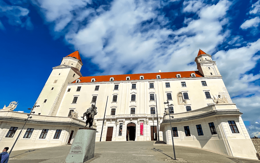 The front of Bratislava castle on a stop during one day in Bratislava. The building is white with a red roof and tower on either side. There is a statue in front of a man on a horse.