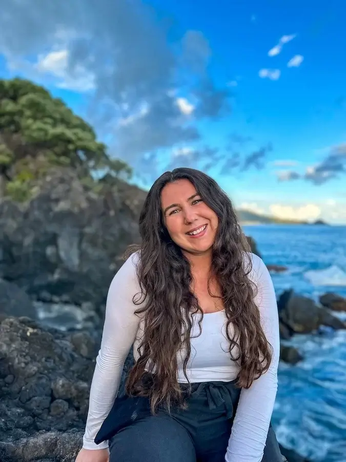 Elaina wearing a white long-sleeved shirt and black pants sitting on a rock next to the ocean in the Azores