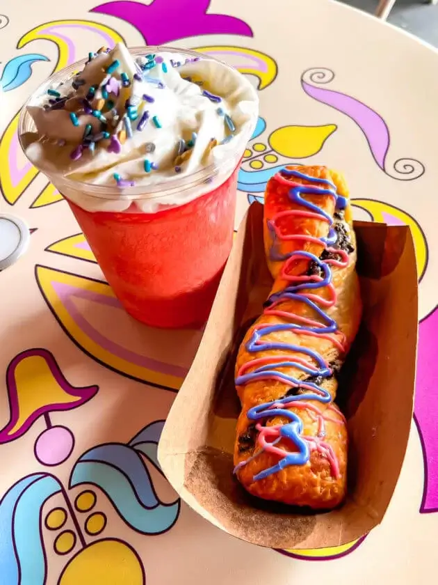 Cheshire cat tail made up of a croissant filled with cream and chocolate and topped with colorful frosting and pink slushie drink with whipped cream and sprinkles
