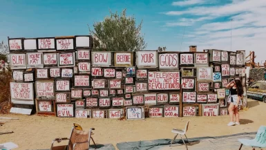 Elaina standing next to the TV Wall art installation in Slab City. The art installation consists of several televisions reading messages about how media influences society.