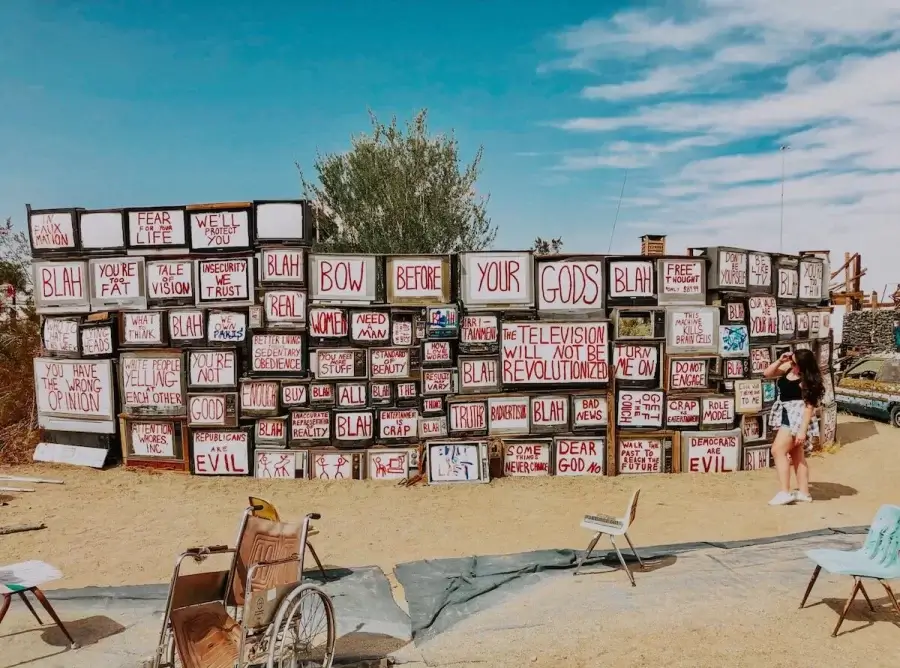 Elaina standing next to the TV Wall art installation in Slab City. The art installation consists of several televisions reading messages about how media influences society.